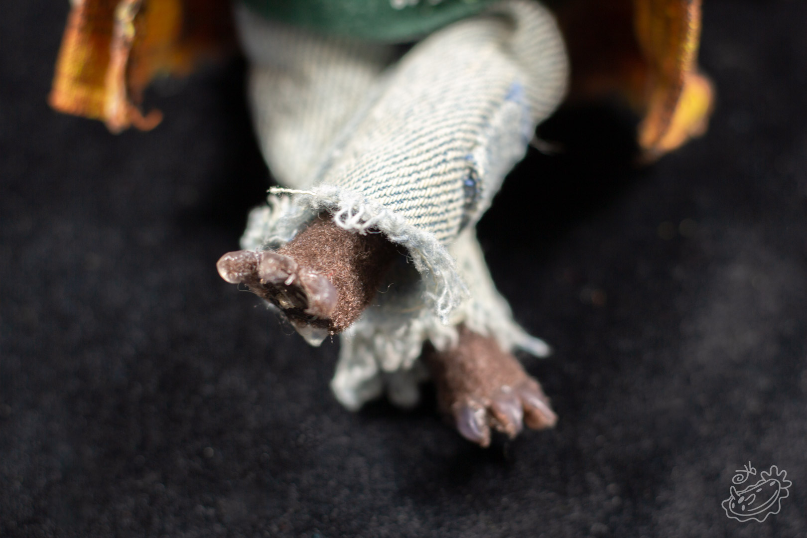 A felt doll of the Michigan Dogman. It is dressed in jeans, a band tank top with the art of the dogman on it, and a brown flannel. It is a close view of the paw details.