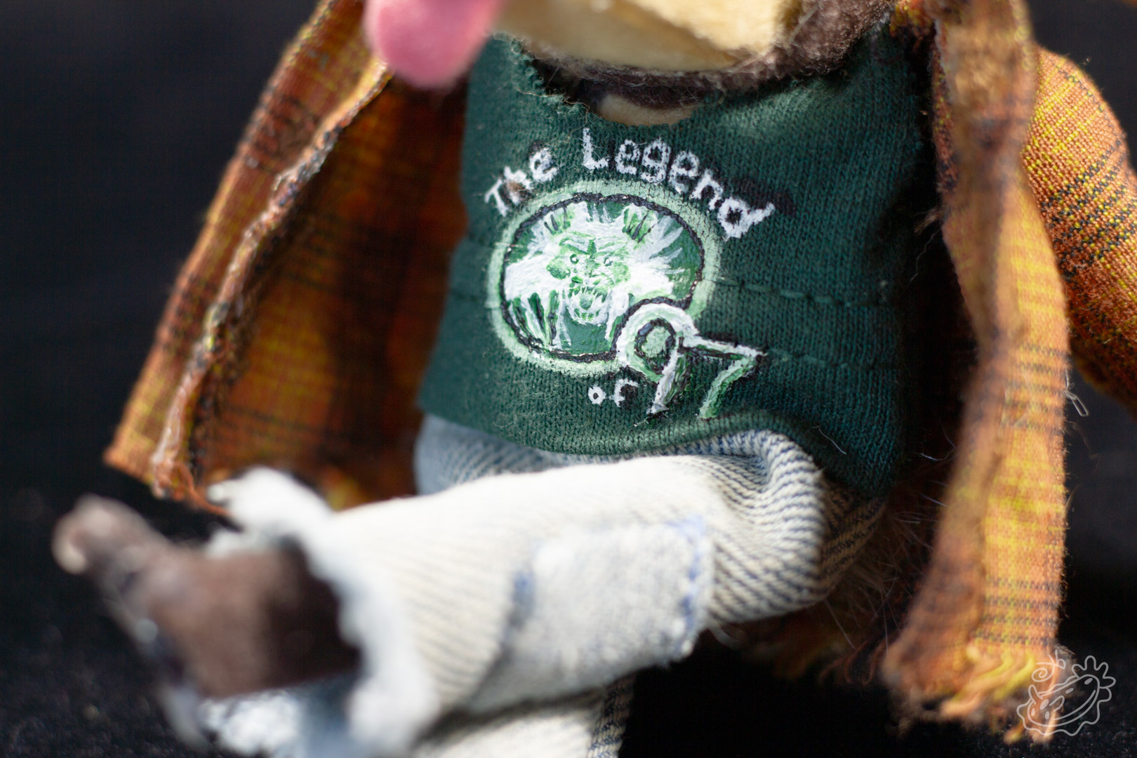 A felt doll of the Michigan Dogman. It is dressed in jeans, a band tank top with the art of the dogman on it, and a brown flannel. It is a close view of the band tank top details.